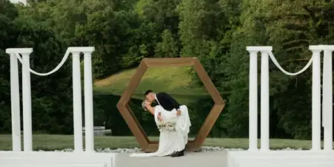 A bride and groom sharing a kiss under a wooden hexagonal arbor at an outdoor wedding venue.