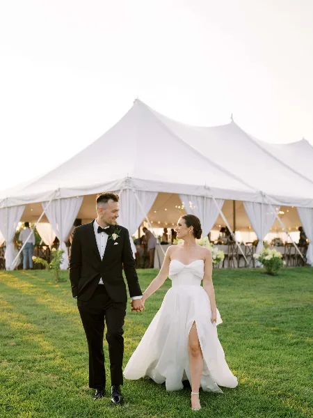 A bride and groom walking hand-in-hand on a lawn in front of a large white wedding tent with visible interior decor.