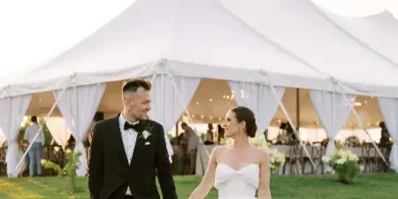 A bride and groom walking hand-in-hand on a lawn in front of a large white wedding tent with visible interior decor.