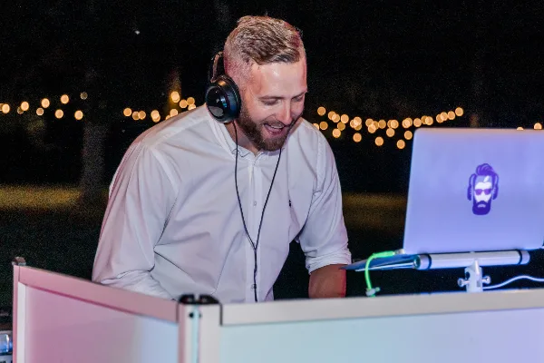 A professional DJ wearing headphones and a white shirt performing at an event with string lights in the background.