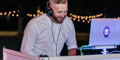 A professional DJ wearing headphones and a white shirt performing at an event with string lights in the background.