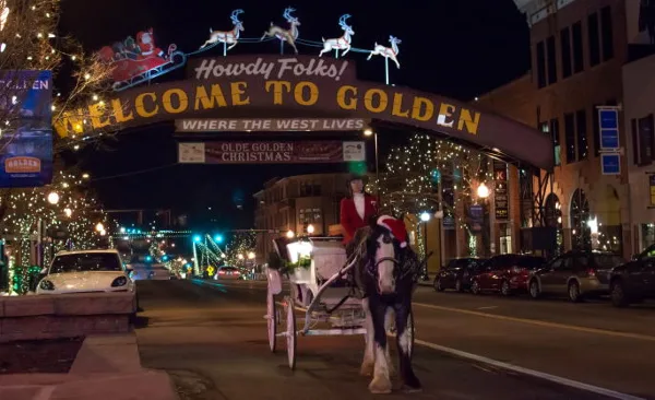 A horse-drawn carriage with a driver in a red coat on a street in Golden, Colorado, near a 'Welcome to Golden' archway.
