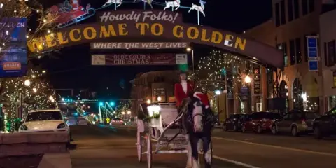A horse-drawn carriage with a driver in a red coat on a street in Golden, Colorado, near a 'Welcome to Golden' archway.