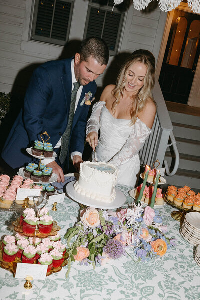 Bride and groom cutting a wedding cake at a decorated table outdoors at a historic mansion venue.