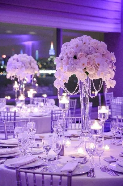 A close-up of an elegant event table setting featuring a tall floral centerpiece with white roses and orchids, surrounded by candles and glassware.