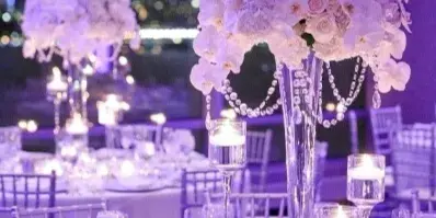 A close-up of an elegant event table setting featuring a tall floral centerpiece with white roses and orchids, surrounded by candles and glassware.