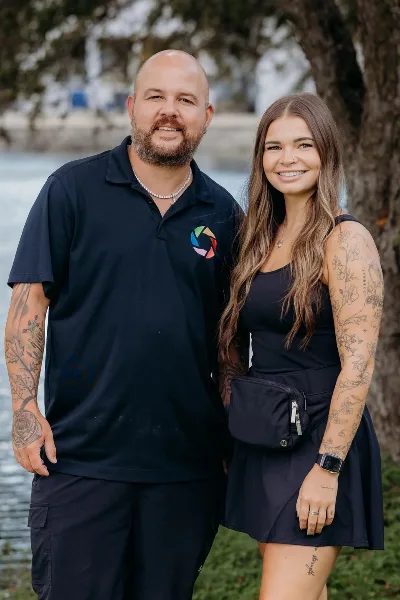 A videographer and a bride posing together outdoors near a body of water.