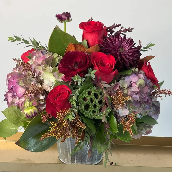 A floral arrangement with red roses, purple hydrangeas, and dark chrysanthemums in a galvanized metal bucket.
