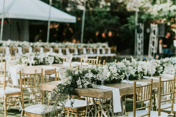 Outdoor event setup featuring long wooden tables, gold Chiavari chairs, and white floral centerpieces.