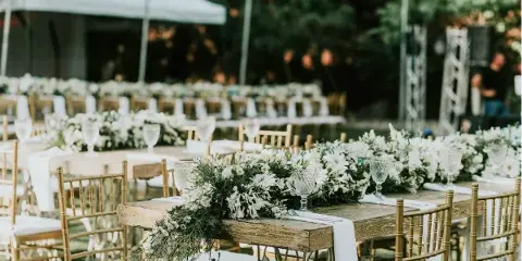 Outdoor event setup featuring long wooden tables, gold Chiavari chairs, and white floral centerpieces.