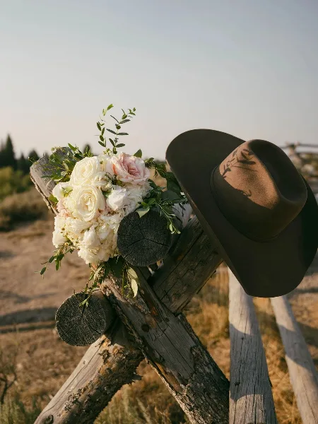 A bouquet of white and pink roses with greenery resting on a rustic wooden fence post next to a brown hat.