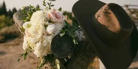 A bouquet of white and pink roses with greenery resting on a rustic wooden fence post next to a brown hat.
