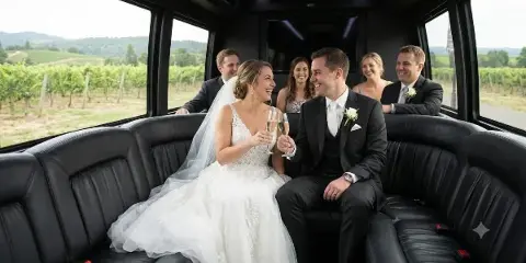 A bride and groom with their wedding party seated inside a luxury charter bus, toasting with champagne.