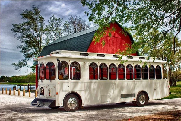 A white trolley bus with red interior curtains parked in front of a red barn.