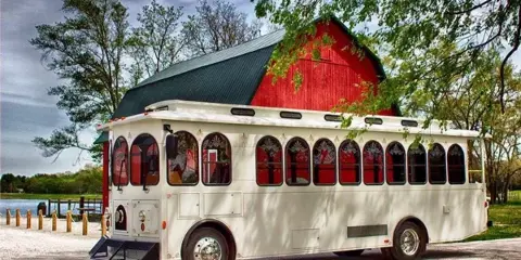 A white trolley bus with red interior curtains parked in front of a red barn.