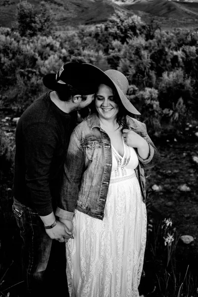 A black and white photograph of a couple standing outdoors in a natural landscape, with the man kissing the woman's forehead.