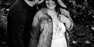 A black and white photograph of a couple standing outdoors in a natural landscape, with the man kissing the woman's forehead.