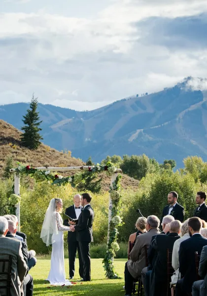 Outdoor wedding ceremony with a floral arch, officiant, bride and groom, and seated guests on a grassy lawn with mountains in the background.