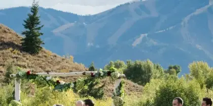 Outdoor wedding ceremony with a floral arch, officiant, bride and groom, and seated guests on a grassy lawn with mountains in the background.