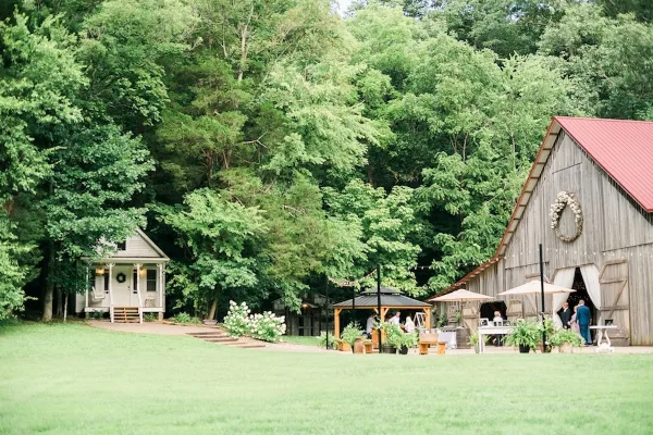 A rustic wooden barn with a red roof and a floral wreath on the facade, situated in a grassy field surrounded by dense green trees.