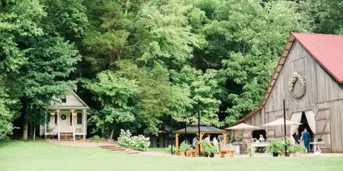 A rustic wooden barn with a red roof and a floral wreath on the facade, situated in a grassy field surrounded by dense green trees.