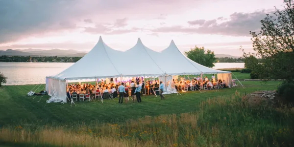 A large white event tent set up on a grassy field near a lake, filled with guests at a wedding reception.