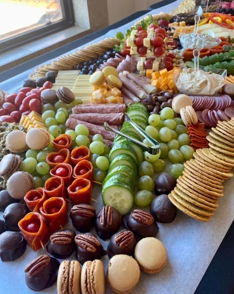 A large, elaborately arranged catering platter featuring sliced meats, cheeses, grapes, cucumber slices, macarons, and chocolate-covered pecans.