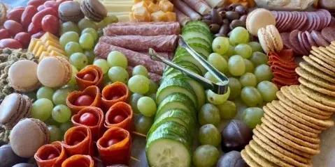 A large, elaborately arranged catering platter featuring sliced meats, cheeses, grapes, cucumber slices, macarons, and chocolate-covered pecans.