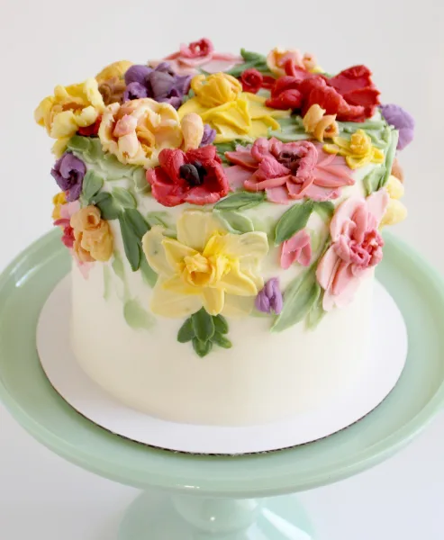 A white frosted cake decorated with colorful hand-piped buttercream flowers on a green cake stand.