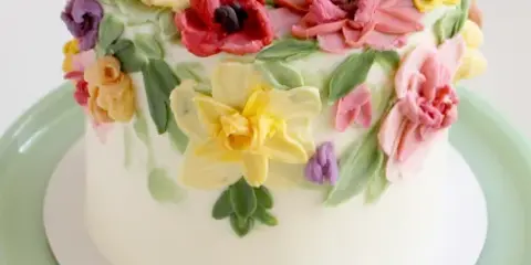 A white frosted cake decorated with colorful hand-piped buttercream flowers on a green cake stand.