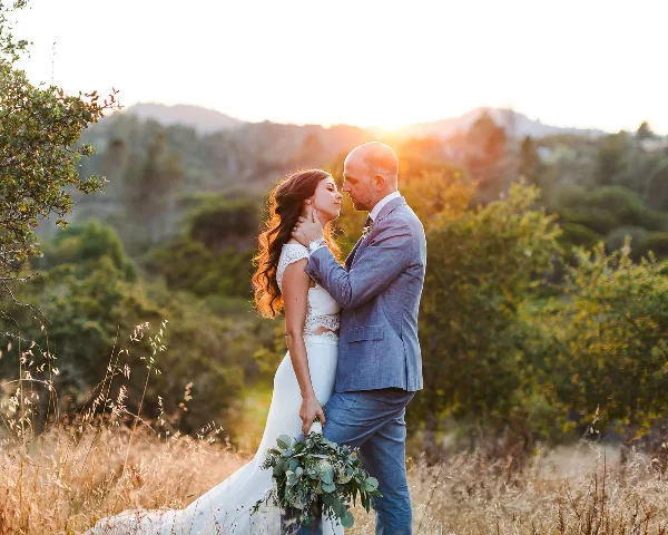 A bride and groom standing close together in a field with a mountainous background at sunset.