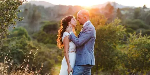 A bride and groom standing close together in a field with a mountainous background at sunset.