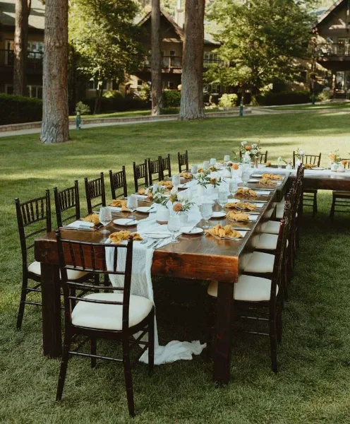 A long rustic wooden dining table with chairs set up on a lawn for an event.