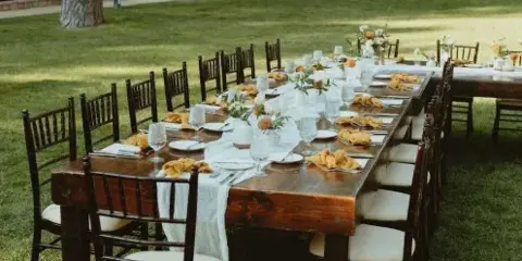 A long rustic wooden dining table with chairs set up on a lawn for an event.