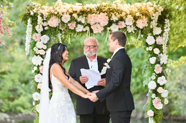 A bride and groom holding hands during a wedding ceremony under a floral arch.