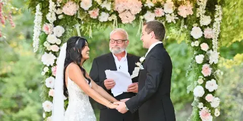 A bride and groom holding hands during a wedding ceremony under a floral arch.