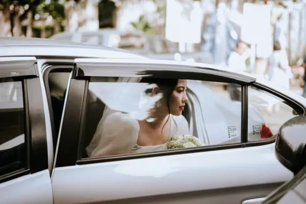 A bride wearing a white wedding dress and veil is seated inside a car, looking out the window.