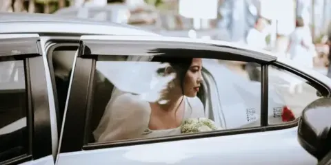 A bride wearing a white wedding dress and veil is seated inside a car, looking out the window.