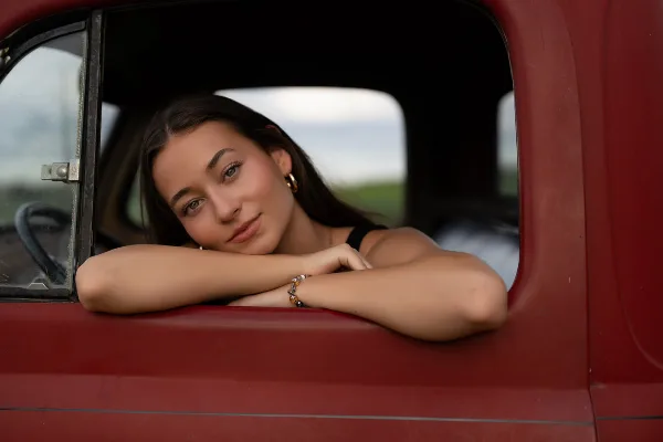 A young woman with dark hair leaning on the window of a red vintage truck, looking at the camera.