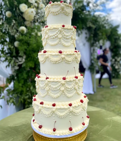 A five-tier white wedding cake decorated with piped lace patterns and red cherries, displayed on a gold stand at an outdoor event.