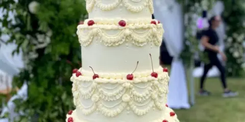 A five-tier white wedding cake decorated with piped lace patterns and red cherries, displayed on a gold stand at an outdoor event.