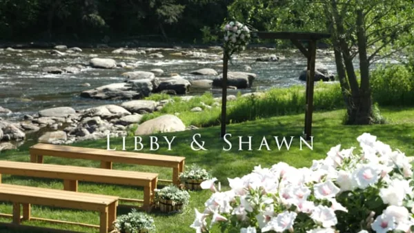 A wedding ceremony setup featuring a floral arch, wooden benches, and a river in the background.