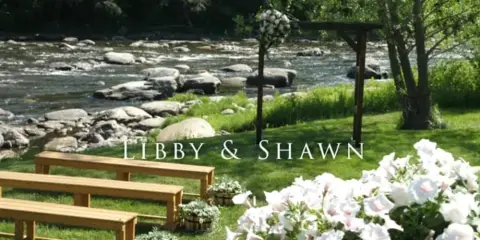 A wedding ceremony setup featuring a floral arch, wooden benches, and a river in the background.