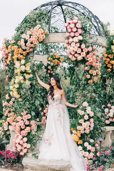 A bride in a floral gown standing inside a large wedding arch covered in lush greenery and cascading roses in shades of peach, yellow, and pink.