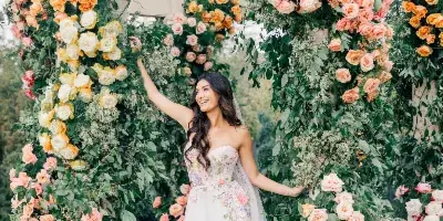 A bride in a floral gown standing inside a large wedding arch covered in lush greenery and cascading roses in shades of peach, yellow, and pink.