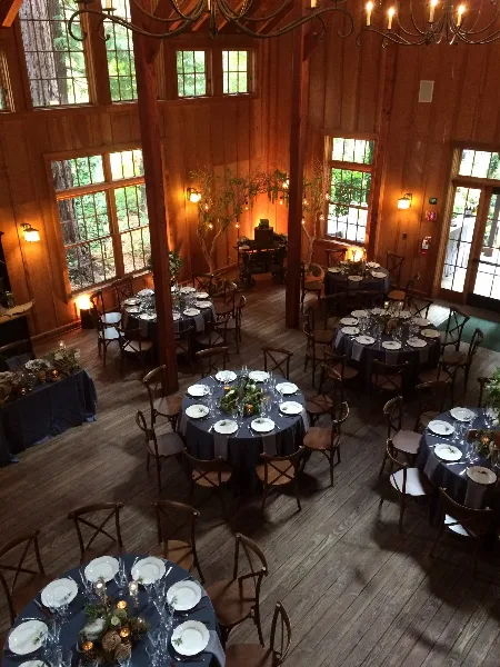 An overhead view of a rustic wooden event hall set up for a wedding reception with round tables, dark linens, and place settings.