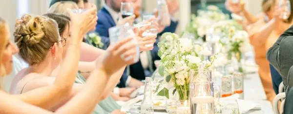 Guests at a wedding reception raising glasses in a toast at a long table decorated with white floral centerpieces.