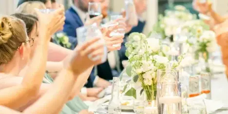 Guests at a wedding reception raising glasses in a toast at a long table decorated with white floral centerpieces.