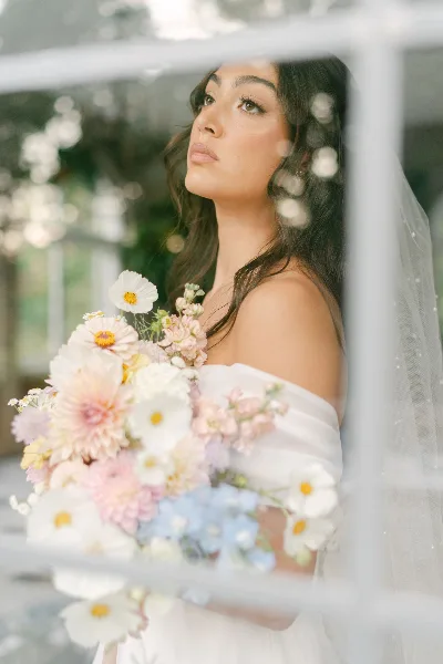 A bride with styled hair and makeup holding a floral bouquet, viewed through a glass pane.