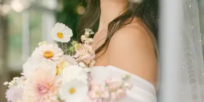 A bride with styled hair and makeup holding a floral bouquet, viewed through a glass pane.